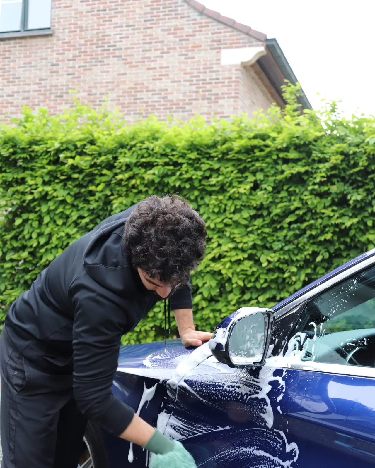 Louay in een zwarte hoodie wast met een groene washand de voorkant van een donkerblauwe auto vol schuim