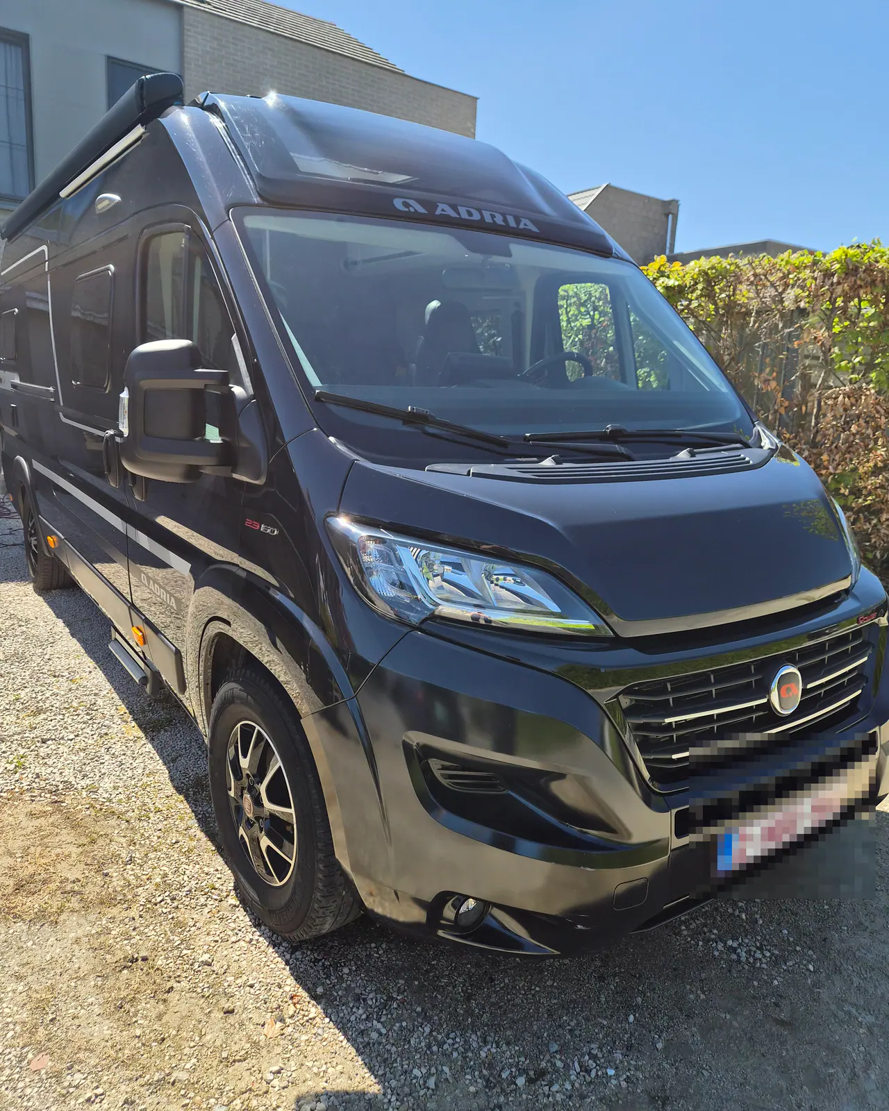 Een zwarte campervan met hoog dak staat op een grindoprit voor een beukenhaag en twee woningen