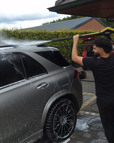 Een man spuit met een hogedrukreiniger water over het dak van een grijze SUV vol schuim