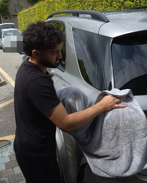 Een man droogt met een grote grijze microvezeldoek de achterkant van een grijze SUV op een oprit