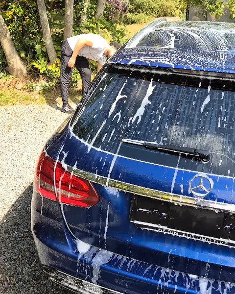 Een blauwe Mercedes-break staat onder wit schuim op een grindoprit, iemand werkt aan de andere zijde