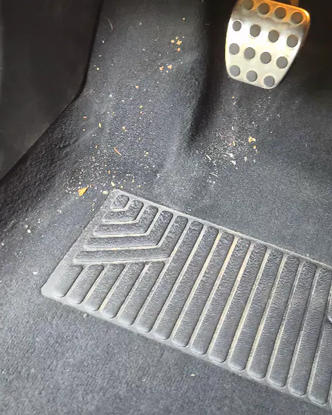 Kruimels en zand op het donkere tapijt van een voetenruimte, naast een geperforeerd metalen pedaal